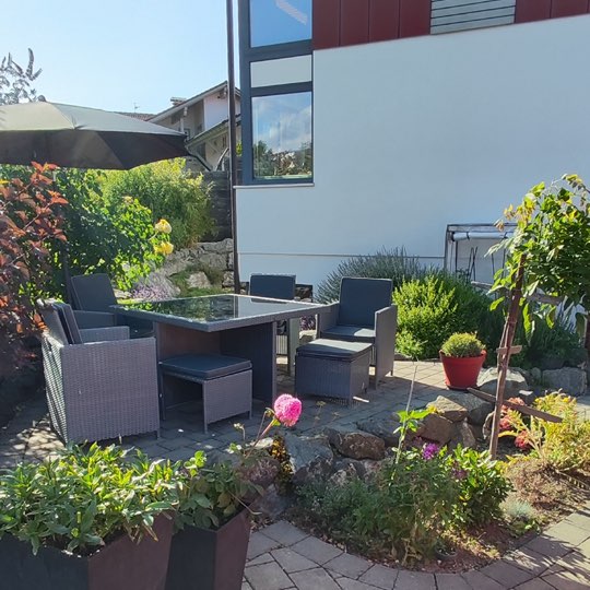Überdachte Sitzecke mit Lounge-Möbeln, Sonnenschirm und blühendem Steingarten vor der Ferienwohnung Alpenglück³