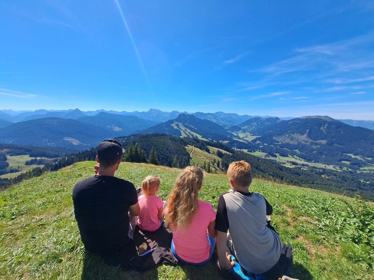 Ein Erwachsener und drei Kinder sitzen von hinten im Gras auf einem Bergrücken und blicken über bewaldete Täler auf die Gipfel der Allgäuer Alpen.